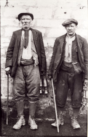 Photograph of two men standing against a light- coloured wall; both men are middle-aged and are dressed in work clothes of heavy boots, gaiters, heavy trousers, waistcoat, jacket and scarf, and carry miners' lamps; both hold sticks in their right hands