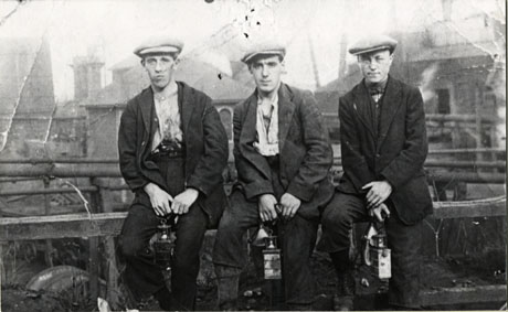 Photograph of three young men sitting on a rail, with indistinct buildings, possibly of the colliery, behind them; they are wearing caps, waistcoats, shirts, scarfs, trousers, gaiters and boots; they are holding miners' lamps; they have been identified as miners from Murton