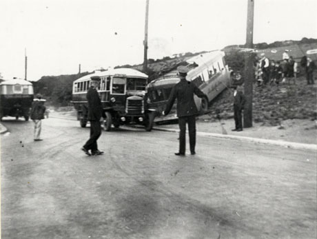 Photograph showing a road running away from the camera, with a motor bus facing the camera, standing stationary in the road on the right; another bus has run up the slope and into a hedge at the side of the road next to the front of the first bus; the back of another bus can be seen facing away from the camera on the left of the road; two policemen with their backs to the camera are standing in the road looking at the bus on the side of the road; a boy in the road and a crowd of people near the hedge can be seen; the photograph has been identified as London Bus Crash At The Water-Works, Murton
