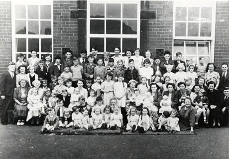Photograph of approximately seventy children, between the ages of approximately six months and fifteen years, posed outside a brick building with large windows accompanied by approximately fifteen adults; the photograph has been identified as Coronation Party, New Hesleden, 2 June 1953