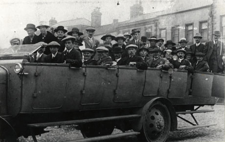 Photograph of a charabanc containing thirty five middle-aged people with the facade of buildings behind them; the charabanc is photographed from the side and the image stretches the width of the photograph; the photograph has been identified as Charabanc Outing, Woods Terrace, Murton, 1920