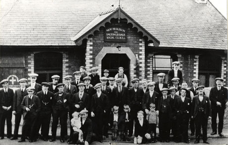 Photograph of a group of thirty one men, eight boys and one infant posed outside a one-storeyed building; above the entrance to the building is a sign reading New Hesledon and Dalton Le Dale Social Club Ltd.