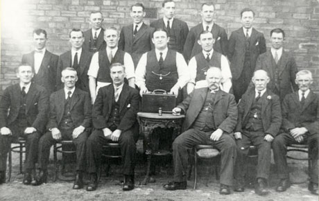 Photograph of seventeen men posed against a wall; the men on the front row are sitting either side of a small iron table on which a man next to it is resting a gavel; three of the men in the middle of the group are in short sleeves; all the rest wear jackets; one of the shirt-sleeved men standing behind the table is resting a small suitcase on the table; the photograph has been identified as Water Works Club Committee
