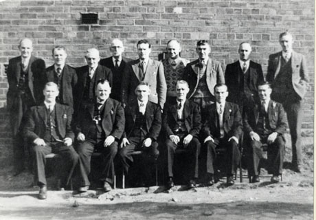 Photograph of fifteen men posed against an exterior brick wall; they are all wearing suits and ties and a number are the same individuals as in murt0018; they have been identified as the Water Works Committee