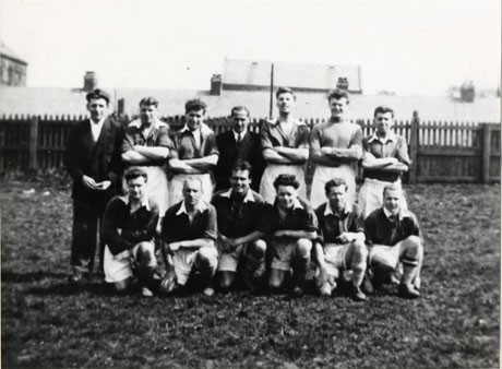 Photograph of eleven men in football strip, accompanied by two other men, posed in a field, with a fence and roofs of low buildings behind them; they have been identified as members of the Water Works Football Club