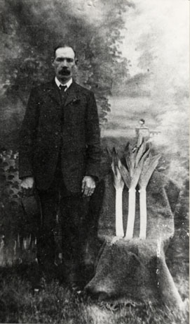 Photograph showing a man full-length, wearing a dark suit and tie, standing beside a chair covered with a cloth on which three leeks are displayed; the photograph has been taken in a photographer's studio; the man has been identified as coming from Cold Hesledon