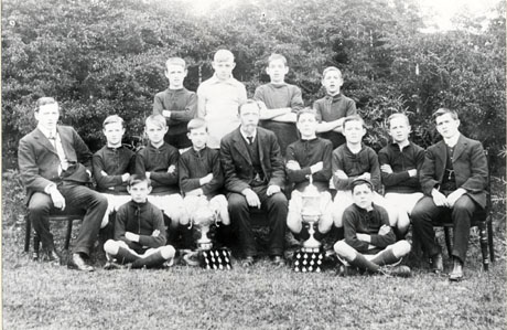 Photograph of twelve boys and three men posed in a field in front of trees;eleven of the boys are wearing football strip and in front of the group are two trophy cups and two boards bearing twelve medals; the boys have been identified as Murton Old School Football Team