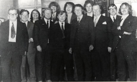 Photograph of twelve men standing in an informal group inside a building, very little of which can be seen; they have been identified as Members at New Hesledon Club, 1972-73