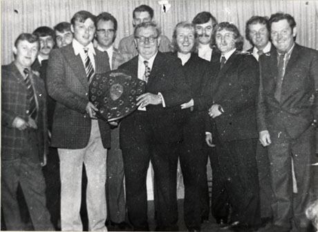Photograph of twelve men in suits standing in an informal group in front of curtains and surrounding two men who are both holding a trophy shield; the shield has been identified as Water Works Darts and Dominoes Murton and District Shield