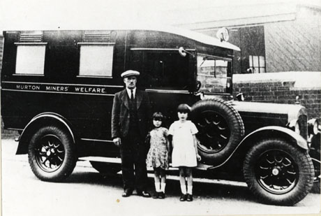 Photograph of the side of a motor van with running board and spare tyre on the running board; the vehicle is photographed on the driver's side and the make and registration number cannot be seen; along the side of the vehicle the words Murton Miners' Welfare are written; in front of the vehicle a middle-aged man and two small girls are standing; the vehicle has been identified as Murton Miners' Welfare Ambulance