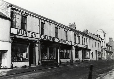Photograph of the exterior of the Murton Colliery Cooperative Society Limited, showing four shop windows, the contents of which cannot be seen clearly; three properties, one of which may be a cinema, can be seen beyond the cooperative store; in the foreground is the surface of the road, which has been identified as Woods Terrace