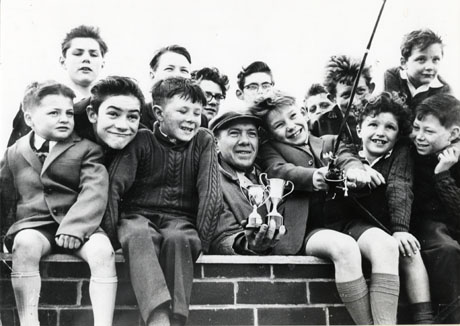 Photograph described as Eight Year Old Boy of Murton Wins Trophy For Fishing, showing four boys sitting on a wall, and the heads of eight boys behind them, looking at a fifth boy sitting on the wall who is holding a fishing rod; immediately on the boy's right is the head and shoulders of a man who is holding two trophy cups
