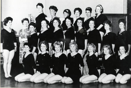 Photograph of twenty one women in leotards, posed against an interior wall, possibly that of a gymnasium; a woman, also in a leotard, is standing at the left of the women and another woman, dressed in a frock, is also standing to the left of the group; they have been identified as Murton Ladies at Keep-Fit Class