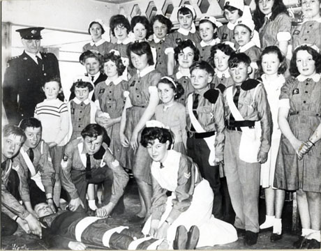 Photograph of eighteen girls and three boys in the uniform of the St. John Ambulance Brigade, watching three boys and one girl in the uniform of the Brigade practising strapping up a casualty; a man in the uniform of the Brigade is standing on the left of the photograph watching the proceedings; the photograph has been identified as St. John Ambulance Cadets, Murton, 1963