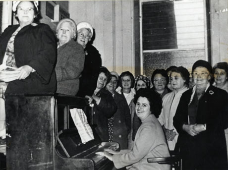 Photograph of fourteen members of Murton Womens' Institute inside an unidentified building; in the middle of the photograph a woman is sitting at an upright piano, playing; round her ten women wearing coats and hats are standing; three more women are sitting on the top of the piano
