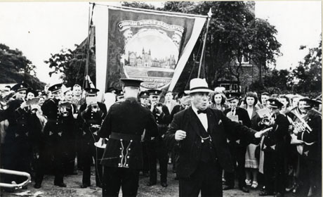 Photograph of Murton Lodge banner, with a picture of Conishead Priory Convalescent Home, being held in front of trees and a building behind the trees; the banner is surrounded by a crowd of people and is preceded by bandsmen in uniform; a man in a suit, bow tie and Trilby hat is standing in the middle of the foreground of the picture; the photograph has been described as Nichol Chapman Leading Murton Banner at Durham