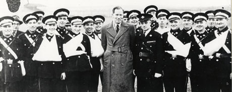 Photograph of nineteen men in uniform, five of whom have arms in slings, standing on either side of a man wearing an overcoat; next to him is a higher ranking officer; the photograph has been identified as Murton and Hetton Ambulance Team