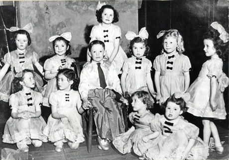 Photograph of ten small girls aged approximately six years dressed alike in short frocks with three bows on the bodice and a large bow in their hair; in the middle of the group of girls is a small boy of approximately the same age in satin tousers and shirt and tie, sitting on a chair; the group has been identified as Tap Dancing Class, Murton Colliery