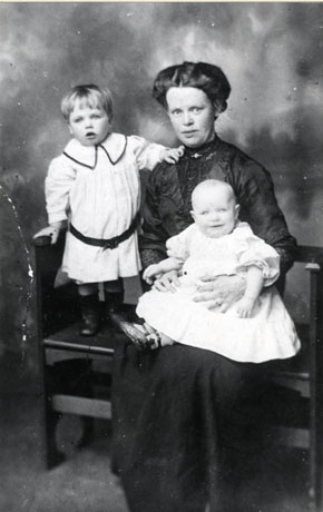 Photograph of a woman sitting on a bench with an infant on her lap and a small boy standing on the bench beside her; she is dressed formally in a dark dress with a brooch at her throat; the infant is wearing an elaborate gown and the small child a smart dress with a belt