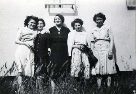 Photograph of one middle-aged and four young women standing in a row in front of a light-coloured wall with long grasses in front of them; they are wearing light summer dresses and appear to be in sunlight; they have been identified as Murton Ladies at Crimdon