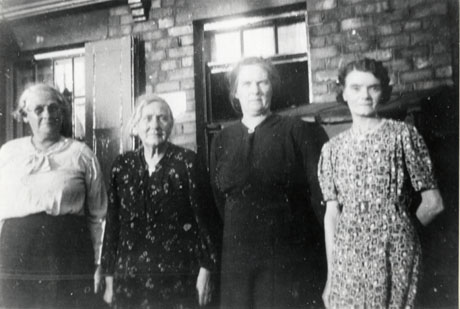 Photograph of four middle-aged women standing in a row in front of a brick building with windows with shutters; three are dressed in light dresses and the fourth is wearing a blouse and skirt; they are described as Four Ladies of Cold Hesledon