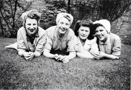 Photograph of the heads and shoulders of four young women lying on grass with a wall and bushes behind them; they are wearing overalls and turbans and have been described as Murton Canteen Staff