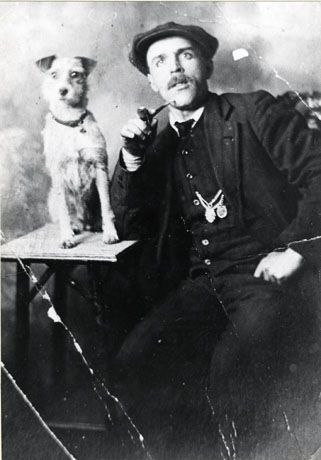 Photograph of a middle-aged man wearing a suit, watch chain and cap, and holding a pipe, seated on the right of the picture; next to him, on the left of the picture, is a small terrier dog standing on a small table; the photograph appears to have been taken in a photographer's studio; they have been identified as Tim Downing with Katy, the Dog, Murton Colliery