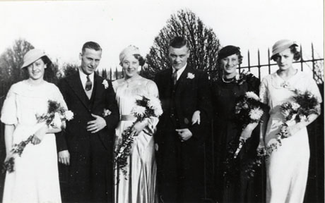 Photograph of four women in formal gowns, carrying bouquets of flowers and wearing hats, standing in a row with two men formally dressed in suits and ties and buttonholes; they are posed against a fence and trees; they have been identified as Wedding, Cold Hesleden, 1937