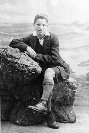 Photograph of a young boy of approximately fifteen years sitting on an artificial rock in a photographer's studio; he is wearing an open-necked shirt, jacket, short trousers, long socks and boots; he has been identified as Mr. George Sinclair, Assistant Secretary of Democratic Club