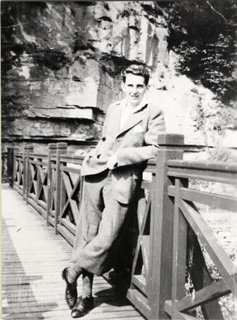 Photograph of a young man standing leaning on the rail of a wooden bridge with a rock face behind him; he is dressed in an open-necked shirt and plus fours; he has been identified as Councillor Short, 1930s