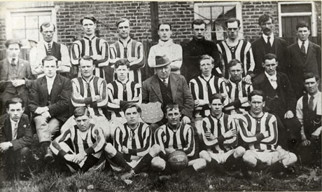 Photograph of eleven members of Murton Villa Athletic Football Club in the club strip, posed with eleven other men in front of a brick building; a man sitting in the middle of the picture is holding a shield with medals on it and a man on the front row is holding a football with the name of the team and the date, 1920-21, on it