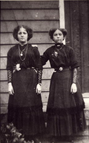 Photograph of two young women posed outside a wooden building, wearing a similar costume of dark high-necked blouse with lace sleeves below the elbow, long two-tiered skirt, chain round the neck and flower attached to the blouse; they have been identified as Mrs. Cowley and Mrs. Neasham