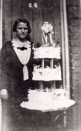 Photograph showing a middle-aged woman standing in a doorway holding a three-tiered wedding cake in her left hand; the door behind her bears the number 25 and she is wearing a dark dress with a light garment beneath it at the neck; she has been identified as Mrs. Evelyn Starkey of Cold Hesledon