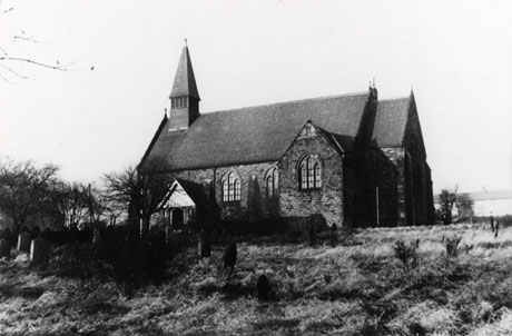 Photograph of the church of Holy Trinity, Murton, taken from the south-east across the graveyard; part of the graveyard and grave stones can be seen; beyond the church, a row of terraced houses can be seen very indistinctly