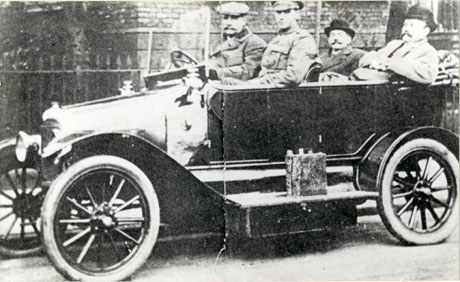 Photograph of an open car with a running board and hard tyres, photographed from the side, with four passengers; in the back, two men with moustaches and wearing bowler hats and overcoats are sitting; in the passenger seat in the front is a man in military uniform, who has been identified as Sergeant McNally; the driver, who is a middle-aged man wearing a heavy coat and a cap has been identified as S. Wood, Colliery Manager; behind the car, part of the wall of a building and some railings can be seen