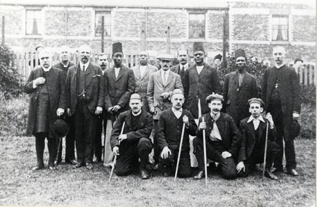 Photograph, described as King of Uganda's Visit to Murton, 1912, showing a group of men posed on a lawn in front of a fence and a terrace of houses; ten men are standing in the group, including three clergymen, one of whom is a bishop; four Africans, three of whom are wearing tall fezzes; and four other Englishmen; on the ground, in front of the men standing, are four men kneeling and holding staves