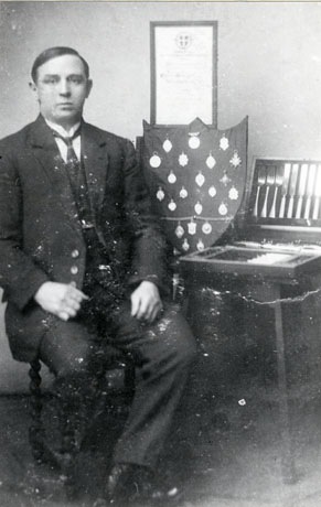 Photograph of a man in a suit and tie sitting on a chair on the left of the picture; on the right of the picture are a table on which a canteen of cutlery is displayed, a shield on which twenty two medals are displayed, and a certificate which is hanging on the wall behind the shield; the man has been identified as Mr. Salkeld, Murton Strong-Man