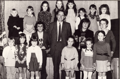 Photograph of a group of seventeen children, and a middle-aged man, posed in front of, and on, a stage, presumably in the hall of a school; fourteen of the children are girls of approximately eight years; the three boys are approximately fourteen years and two of the three can be seen holding trophy cups;the photograph has been identified as recording a Road Safety Presentation 1974