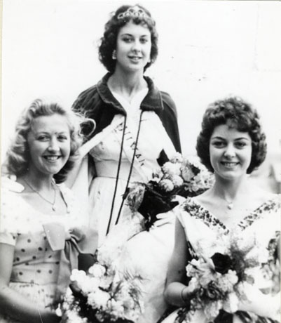 Photograph of the head and torso of three young women wearing summer dresses; the woman in the centre is standing and is wearing a cloak and a tiara and is carrying a bouquet of flowers; the other two are sitting to either side of her and are carrying bouquets of flowers; they are described as Murton Beauty Queen with Attendants