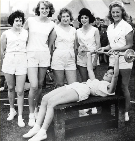 Photograph of five young women dressed in shorts and Plimsolls standing in a row watching a sixth young woman who is lying on her back on a bench holding up a weight; behind the women four men can be seen indistinctly watching the women; the photograph has been identified as Members of Murton Weight-Lifting Club, 1960; the individuals have been identified as, left to right: Margaret Wright; Edna Longstaff; Shiela Miller; Judy Loftus; Joan Little