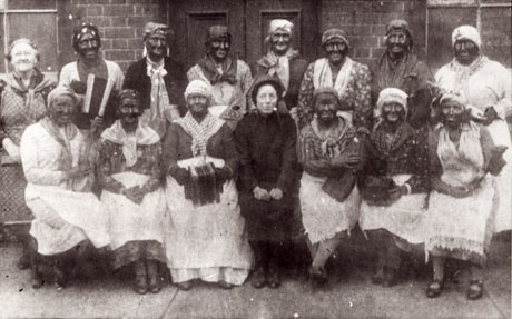 Photograph described as Old Salvation Army- Mrs. Sugden and Mrs. Annie Coxon, showing a group of thirteen people dressed in skirts, blouses, turbans, shawls and aprons and with their faces blacked; six of them are holding musical instruments; at the left end of the back row is a woman wearing an apron and no blacking on her face; in the middle of the front row is a woman dressed in the uniform of the Salvation Army