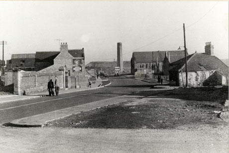 Photograph of a street in Murton,showing the road curving away from the camera to the right; in the foreground on the right is an area of open ground where a building has been demolished; behind that is a low building and behind that a church, identified as the Wesleyan church; beyond the church is a chimney, identified as Coke Chimney and beyond that in the distance a spoil heap; next to the chimney a double decker bus can be seen; on the opposite side of the road the side of a building can be seen with an advertisement for Players' Digger (cigarettes or tobacco); two men can be seen with their backs to the camera