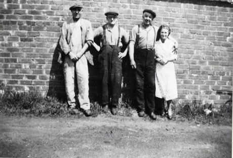 Photograph of three men and a woman standing with their backs to a wall in sunlight; the three men are wearing caps and working clothes and the woman is wearing an overall; they have been identified as Three Baths Attendants and Canteen Assistant, Murton Colliery