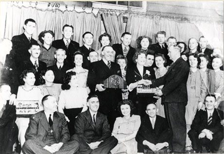 Photograph of thirty five members of the Murton Amateur Operatic Society, in ordinary formal dress, witnessing a presentation of a clock to Mr. Rundell and an object to woman standing next to the recipient of the clock and presumably his wife; Mr. Rundell is holding the clock and his wife is in the act of being presented with her gift by a middle-aged man