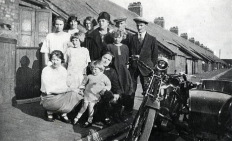 Photograph of a group of two men, six women and two children outside a house in St. Andrew's Terrace, Cold Hesledon; one side of the street showing the backs of the houses can be seen behind the group; next to the group at the right of the picture a motor cycle and side car can be seen