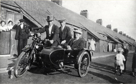 Photograph of the backs of the houses on one side of St. Andrews Terrace, Cold Hesledon, looking down the street, as in murt0077; two women as in murt0077 can be seen standing behind the fence of a house, against which a man is leaning; two men are riding a motor cycle, registration PT 3116, and a man is sitting in the side car;four children can be seen playing in the street