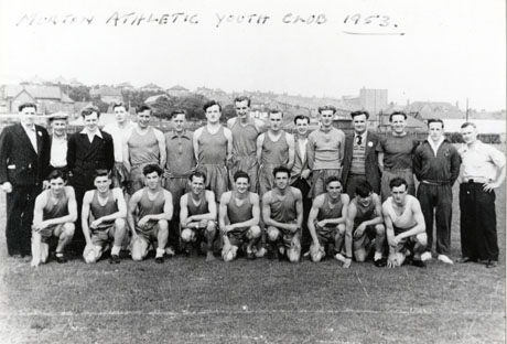 Photograph of thirteen young men in singlets and shorts, posed with five men in track suits and six men in suits, in the open air, with a village, presumably Murton, in the distance behind them; the photograph has the following written on it: Murton Athletic Youth Club 1953