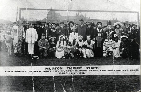 Photograph of approximately a hundred people grouped on a field with a large building very indistinctly in the distance; the figures at the front of the group are in Fancy dress, dressed as clowns, pierrots, soldiers, Mexicans and cowboys; there appear to be goal posts behind the group and the photograph has a printed caption as follows: Murton Empire Staff Aged Miners' Benefit Match By Murton Empire Staff and Waterworks Club, March 21st 1913