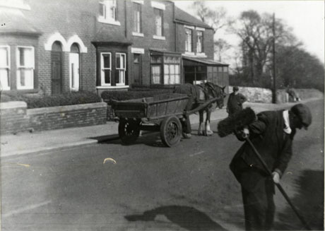 Photograph of four terraced houses at the end of a terrace, with a wall round woods further along the road; in front of the houses, a horse and cart is standing; a man is standing near the horse's head and another man can be seen in the foreground of the right of the picture stooping with a broom; the face of neither man can be seen; the photograph has been identified as follows: Vicarage Terrace, Durham Road(Showing Council Workers)