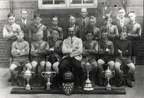 Photograph of eleven boys in football strip, two boys in ordinary clothes, and three men, posed outside a brick building; in front of the boys are four trophy cups and a shield on which there are medals; in front of the man on the front row is a football with the words Murton 1938-39 Council (School)